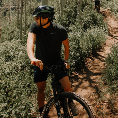 A male mountain biker pauses on his bike to look right at the terrain of a heavily-forested area. Image source: Unsplash.